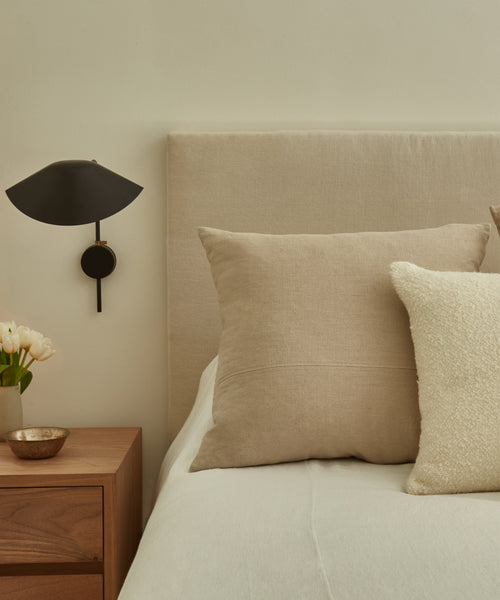 A neatly made bed with a Linen Pillow and light blanket sits beside a wooden nightstand holding a small bowl, white flowers in a vase, and a modern black wall sconce, giving the room a calm, minimalist style.