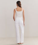 A woman faces away with her hands in her pockets, wearing a white sleeveless ribbed top and Riley Trouser high-waisted relaxed fit summer trousers against a plain light background.