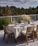 An outdoor dining table set with eight wooden chairs, plates, glasses, a vase of white flowers, and a Linen Tablecloth on a patio overlooking greenery beneath a partly cloudy sky.