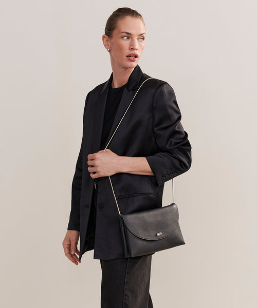 A person poses against a plain light background, wearing a black blazer, black shirt, and jeans while holding the Loren Clutch Bag.