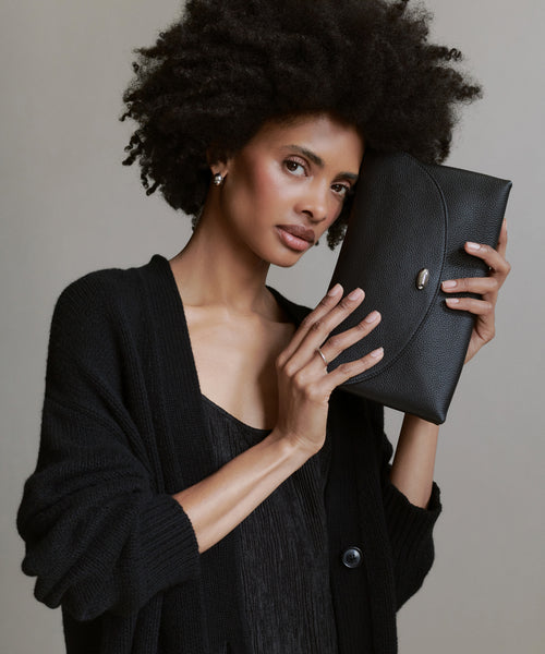 A woman with natural curly hair, dressed in black, holds the Loren Clutch Bag—a striking black leather accessory—next to her face, looking confidently at the camera against a neutral background.