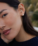 Close-up of a woman wearing Large Loren Pearl Studs—single round freshwater pearl earrings. She has smooth skin, long dark hair, a dark knitted sweater, and the blurred background is green foliage.