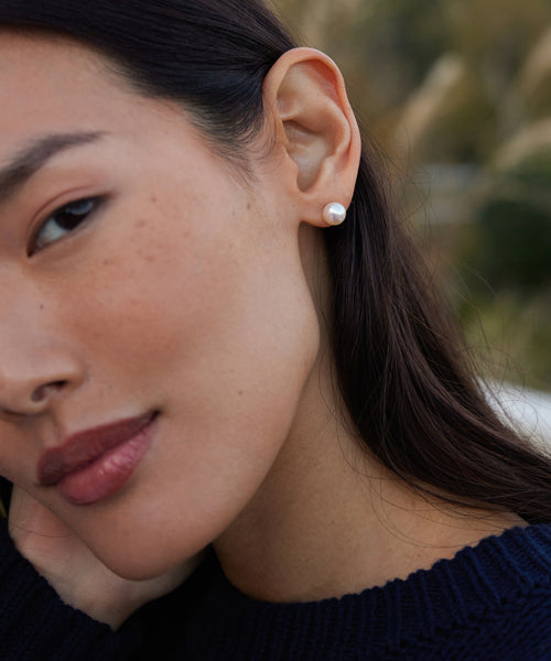 Close-up of a woman wearing Large Loren Pearl Studs—single round freshwater pearl earrings. She has smooth skin, long dark hair, a dark knitted sweater, and the blurred background is green foliage.