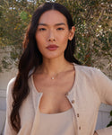 A woman with long dark hair and neutral makeup stands outdoors among greenery, wearing a light beige cardigan, a matching top, and the Loren Pearl Ear Cuff. She looks confidently at the camera.