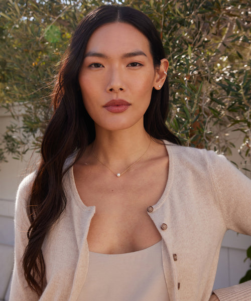 A woman with long dark hair and neutral makeup stands outdoors among greenery, wearing a light beige cardigan, a matching top, and the Loren Pearl Ear Cuff. She looks confidently at the camera.