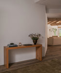 A minimalist light brown wooden console table styled with books, a small candle, and a dark metal vase of dried flowers, set against a white wall in a modern home interior.