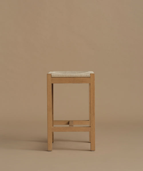 A wooden bar stool with a light beige woven seat cushion, shown against a solid warm beige background.
