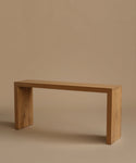 A minimalist light oak wood entry table with a rectangular top and blocky legs, photographed against a solid warm beige background.