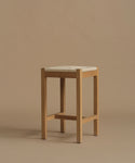 A minimalist wooden stool with a light natural oak frame and a square woven beige seat cushion, set against a solid warm beige background.
