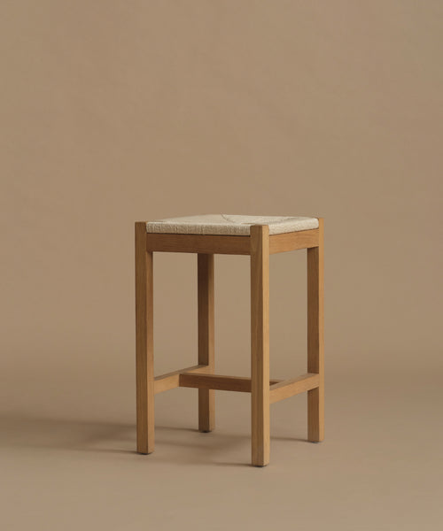 A minimalist wooden stool with a light natural oak frame and a square woven beige seat cushion, set against a solid warm beige background.
