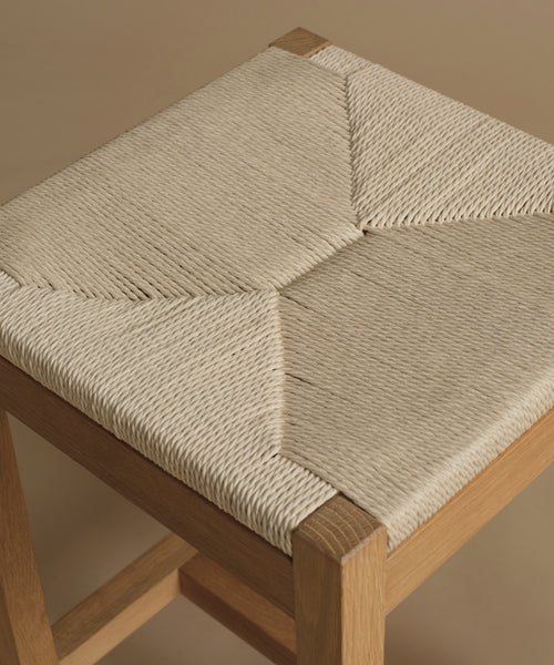 Close-up of a wooden stool with a woven rope seat in a light beige color, set against a solid warm beige background.