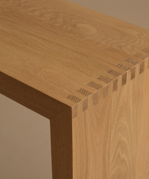 Close-up of a light oak wood table corner showing the wood grain and dovetail joint construction.
