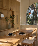 A warm, rustic kitchen interior featuring light natural wood cabinetry, a matching wooden dining table, and a large arched window looking out onto trees.