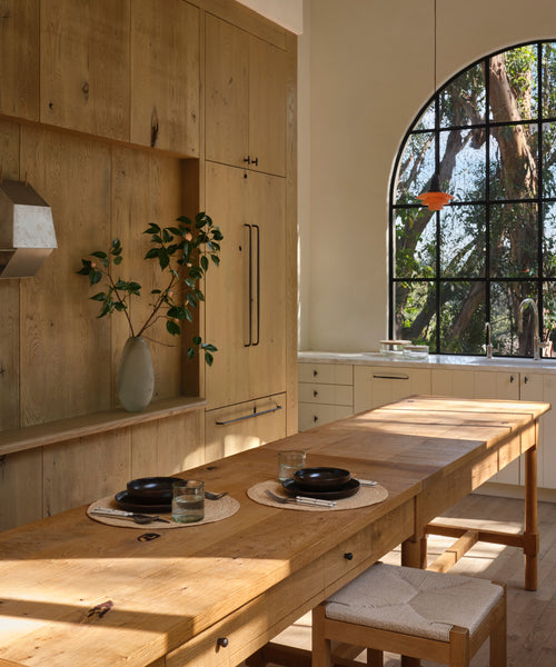 A warm, rustic kitchen interior featuring light natural wood cabinetry, a matching wooden dining table, and a large arched window looking out onto trees.