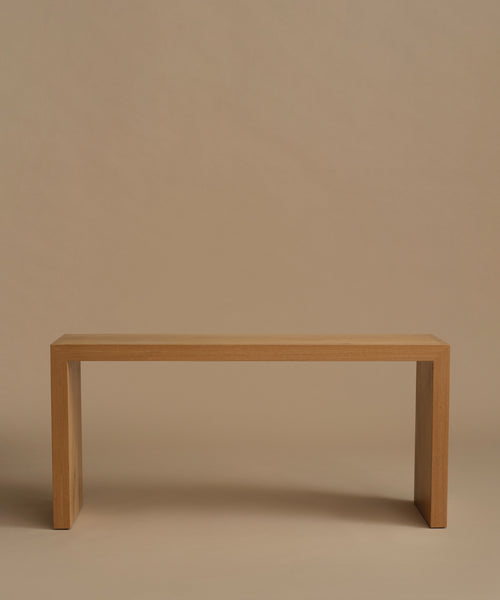 A minimalist light oak wood entry table with a rectangular top and blocky legs, photographed against a solid warm beige background.