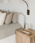 A minimalist bedroom with a white bed, beige and taupe pillows, a Malibu Side Table in light wood as the nightstand, a small ceramic vase, a wooden tray, and a black wall-mounted reading light.