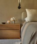 A cozy bedroom features a light wood nightstand with a Mandeville Glass Candle, a bowl, and white flowers next to a bed with beige bedding, all set against a neutral wall and illuminated by a wall sconce above.