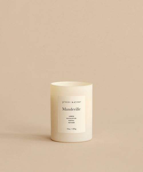 A white Mandeville Glass Candle in a minimalist container rests on a beige surface. Its label lists Lemon, Eucalyptus, and Vervain scents and notes that its hand-poured in Los Angeles.