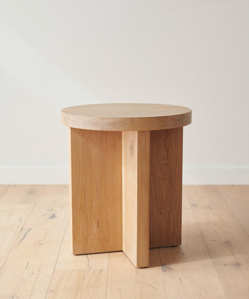 The Marin Side Table, crafted from solid oak, features a round top and geometric intersecting rectangular legs, shown on a light wood floor against a plain white wall.