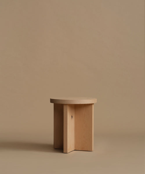 A minimalist light natural wood side table with three intersecting vertical legs, set against a solid warm beige background.