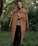 A woman stands on grass, smiling and wearing the McCall Trench Coat over a beige sweater and black pants, with green trees and bushes in the background.