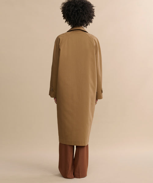 A person with curly hair faces away, wearing the McCall Trench Coat over brown pants, set against a beige background.