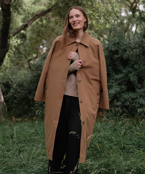 A woman stands on grass, smiling and wearing the McCall Trench Coat over a beige sweater and black pants, with green trees and bushes in the background.