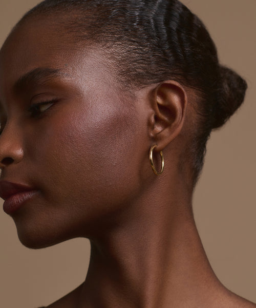 A close-up side profile of a woman with smooth brown skin wears the Medium Eloise Hoops by Jenni Kayne. Her hair is pulled back in a neat bun, and the background is plain beige.
