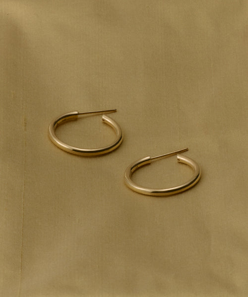 The Medium Eloise Hoops—crafted from 14k gold with straight posts—offer an elegant, heirloom-inspired look, shown here on a light brown fabric background.