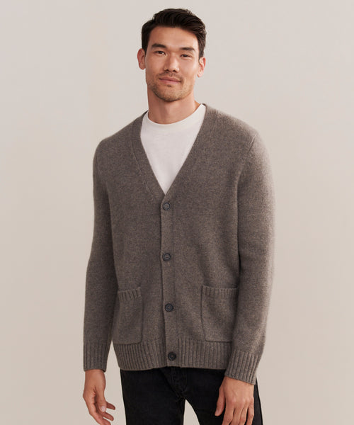 A man stands against a neutral background, wearing a Mens Cashmere Cardigan over a white T-shirt and black pants, his left hand relaxed at his side and a slight smile on his face.