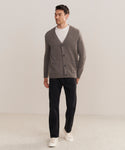 A man stands against a plain light background, wearing a Mens Cashmere Cardigan in gray over a white T-shirt, paired with black pants and white sneakers. He looks to the side with one hand in his pocket.