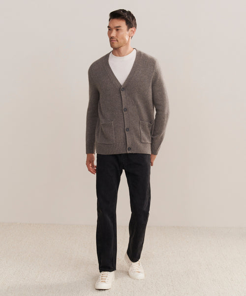 A man stands against a plain light background, wearing a Mens Cashmere Cardigan in gray over a white T-shirt, paired with black pants and white sneakers. He looks to the side with one hand in his pocket.
