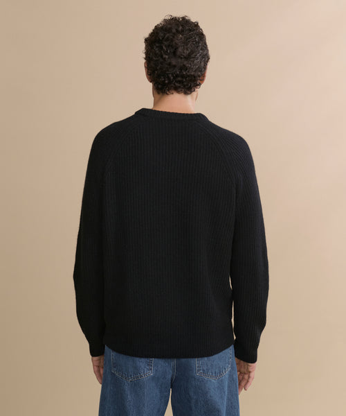 A man with short curly hair, seen from behind, wears the Men's Cashmere Fisherman Sweater in black and blue jeans against a plain beige background.