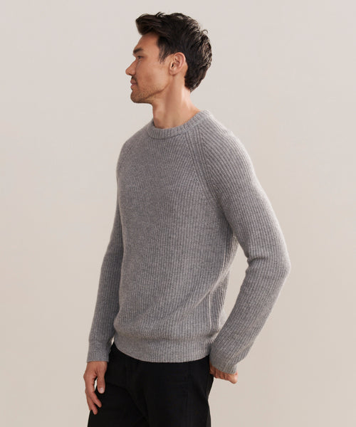A man with short dark hair, dressed in a heather grey Mens Cashmere Fisherman sweater and black pants, stands facing left against a plain light background.