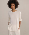 A person with short curly hair stands against a neutral background, wearing an oversized light cream Merino Tee and matching pants, one hand in their pocket.
