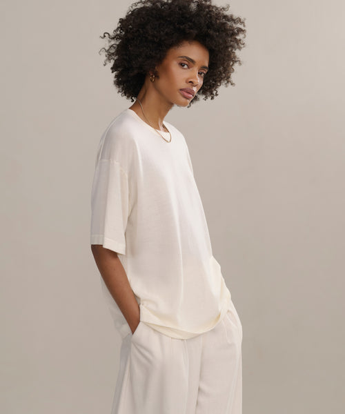A woman with short curly hair stands with her hands in her pockets, wearing the Merino Tee and matching loose-fitting pants—both oversized—set against a neutral background.