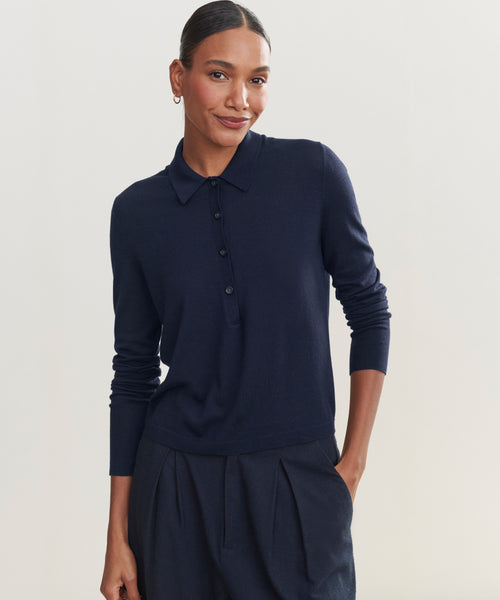 A woman stands against a plain light background, wearing the Grayson Pullover—a navy blue top with a polo placket—paired with matching navy pleated pants. Her hair is pulled back and she smiles softly, one hand in her pocket.
