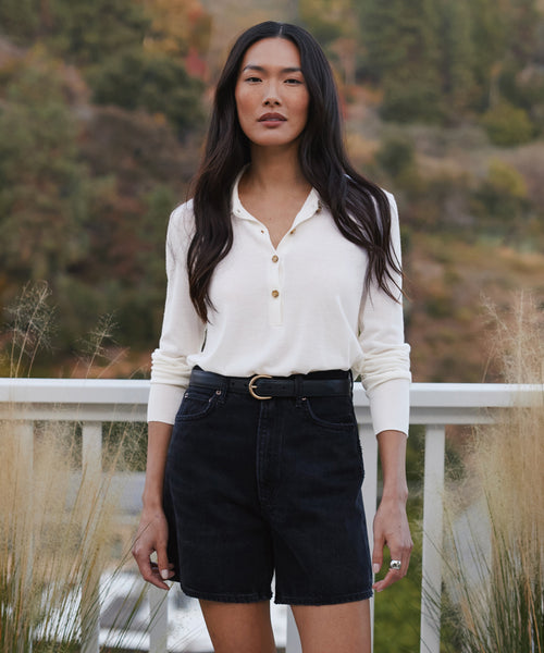 A woman with long dark hair stands outside by a white railing, wearing the Grayson Pullover tucked into belted high-waisted black shorts. Trees and tall grasses in the background complete this perfect warm-weather sweater look.