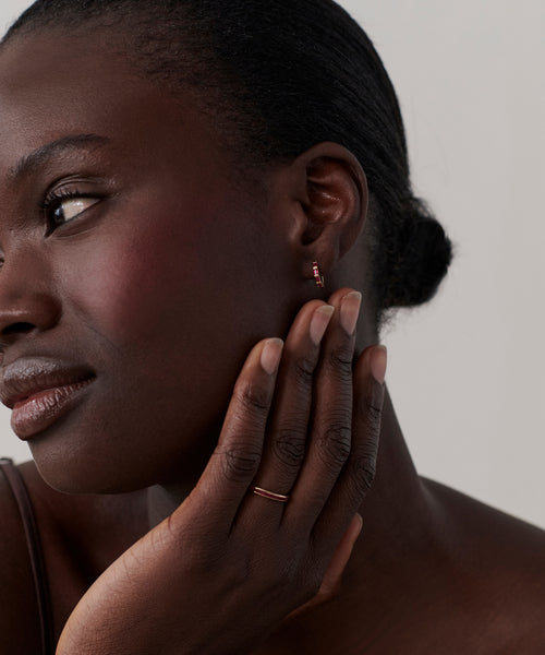 A woman with dark skin and slicked-back hair touches her cheek, showcasing a rose gold ring and elegant Merit Baguette Hoops in 14k solid gold as she gazes thoughtfully to the side against a neutral background.