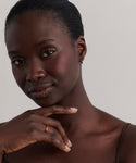 A woman with dark skin and short, pulled-back hair gazes at the camera, her chin resting on her hand. She wears a brown top, a simple ring, and Merit Baguette Hoops against a plain background.