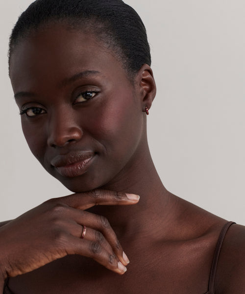 A woman with dark skin and short, pulled-back hair gazes at the camera, her chin resting on her hand. She wears a brown top, a simple ring, and Merit Baguette Hoops against a plain background.
