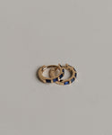 The Merit Baguette Hoops, featuring two small 14k gold hoops with rectangular blue sapphire gemstones, are displayed on a textured light gray surface.