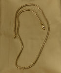 The Merit Chain, a 14k solid gold necklace with a lobster clasp, features medium thickness and a flat, interlocking curb link design. It's showcased on a light tan fabric background.