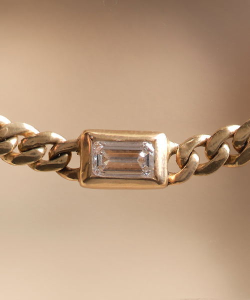 Close-up of the Merit Emerald Cut Necklace, showcasing a rectangular emerald-cut diamond set in a gold bezel, with a softly blurred warm-toned background.