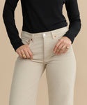 A person in a black long-sleeve top and Mid Rise Wide Leg beige corduroy pants stands against a tan background, hands in their pockets and several gold rings on their fingers.