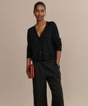 A woman with her hair pulled back smiles gently, standing against a beige background. She wears the Mira Cardigan with loose black pants, gold jewelry, and holds a rust-colored clutch while looking slightly to the side.