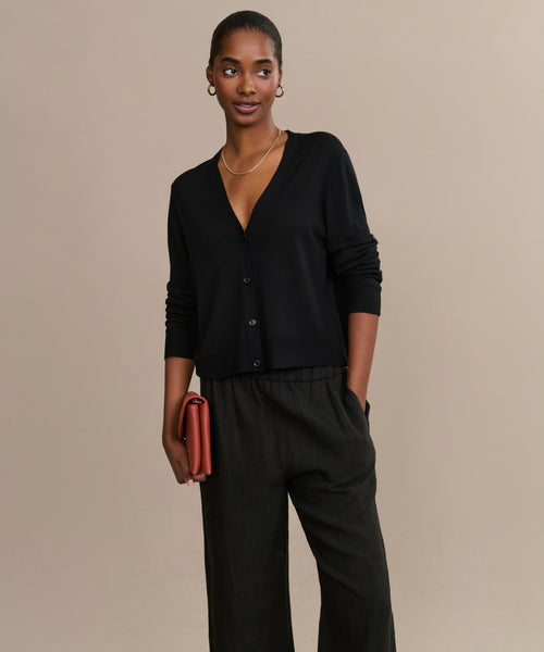 A woman with her hair pulled back smiles gently, standing against a beige background. She wears the Mira Cardigan with loose black pants, gold jewelry, and holds a rust-colored clutch while looking slightly to the side.