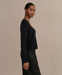 A woman stands in profile against a beige background, wearing the Mira Cardigan with a dark skirt. Her hair is pulled back, and she accessorizes with hoop earrings and a thin necklace, looking at the camera with a slight smile.