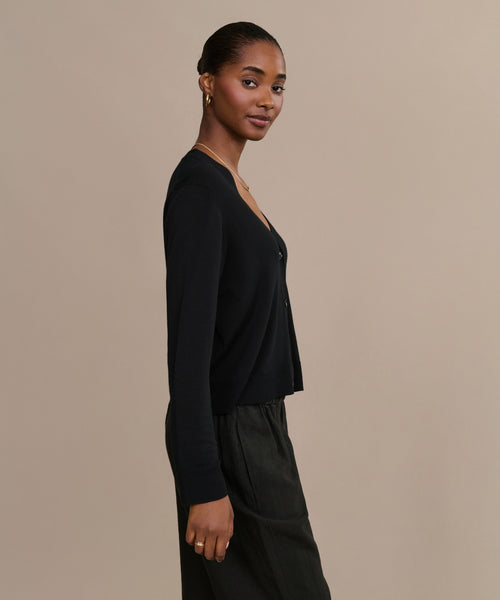 A woman stands in profile against a beige background, wearing the Mira Cardigan with a dark skirt. Her hair is pulled back, and she accessorizes with hoop earrings and a thin necklace, looking at the camera with a slight smile.