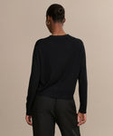 A person with short hair, wearing the black Mira Cardigan and black pants, stands facing away from the camera against a plain beige background.